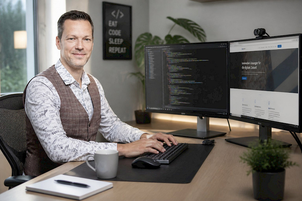 Thomas Wohlmuth lächelnd an seinem Schreibtisch, vor zwei Bildschirmen mit Programmcode und einer Website